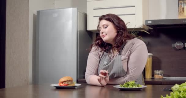Νέοι συν μέγεθος γυναίκα επιλέγοντας μεταξύ πράσινη σαλάτα και burger στην κουζίνα. Διατροφή, βιολογικά τρόφιμα και υγιεινή διατροφή — Αρχείο Βίντεο