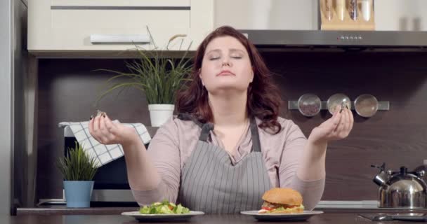 Młoda plus size kobieta medytująca wybierając między zieloną sałatkę i hamburgera, starając się nie jeść fast foodów w kuchni. Dieta, siła woli i koncepcja zdrowego odżywiania — Wideo stockowe