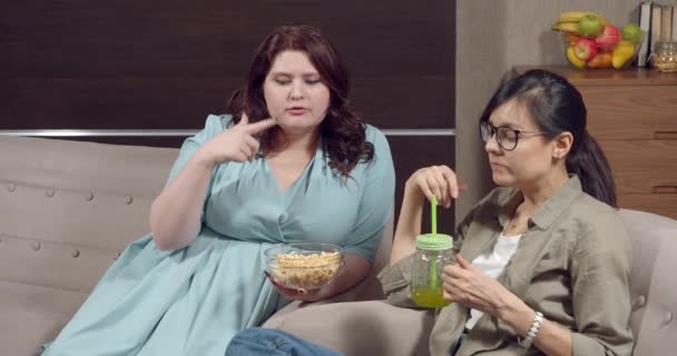 Две молодые женщины лучшие друзья едят попкорн из миски и болтают, сидя дома на диване. Концепция дружбы и общего досуга — стоковое видео