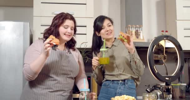 Two cheerful women chatting eating delicious burgers and recording a video for their vlog or leading a live stream on social media using a smartphone in the kitchen — Stock Video