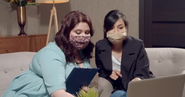 Deux jeunes collègues féminines portant des masques de protection discutent de quelque chose tout en travaillant sur un ordinateur portable dans un bureau moderne — Video
