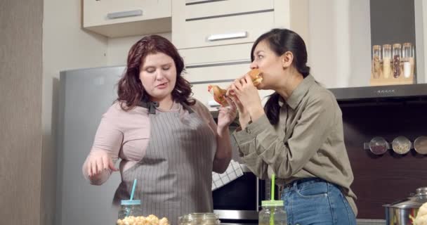 Two young women friends eating juicy burgers and chatting in the kitchen. — Stock Video
