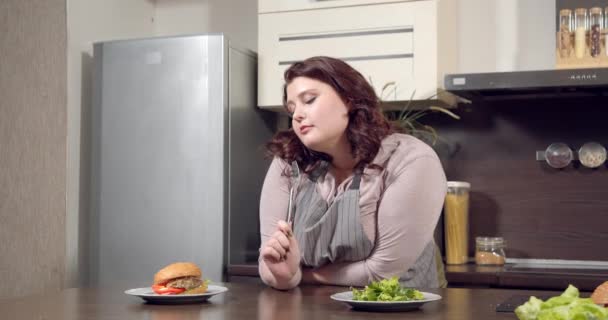 Young plus size woman choosing between green salad and burger in the kitchen. Diet, organic food and healthy eating concept. — Stock Video