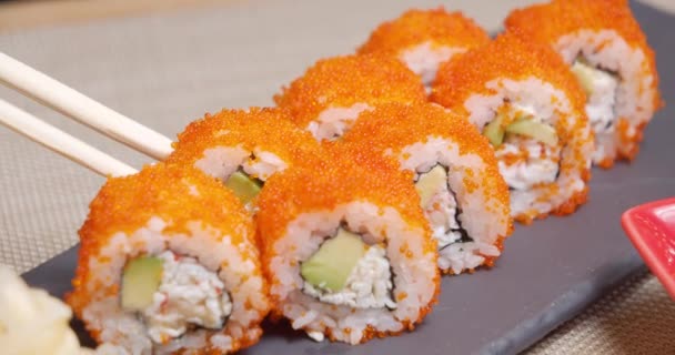 Close-up shot of bamboo chopsticks holding delicious roll with flying fish roe, avocado and soft cheese and dipping into a bowl with soy sauce. — Stockvideo