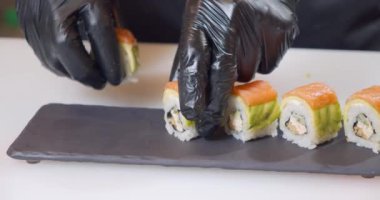 Close-up shot of a chef hands in black gloves serving delicious sushi rolls with avocado and salmon on a plate.
