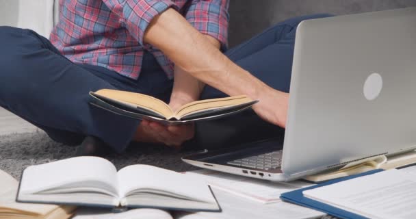 Nerozpoznatelný mladý muž studuje on-line nebo dělá domácí úkoly pomocí notebooku a knihy sedí na podlaze doma — Stock video