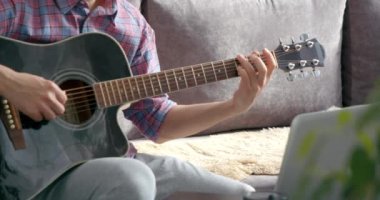 İnternette akustik gitar dersi veren tanınmayan genç adam evdeki kanepede oturup dizüstü bilgisayar kullanıyor.