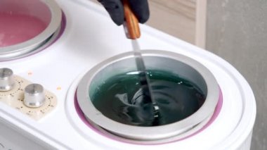 Siyah eldivenli bir güzellik uzmanının özel bir aletle sıcak kozmetik cilasını spatula kullanarak karıştırması. Kaplıcada tüy dökülmeden önce balmumu hazırlama işlemi
