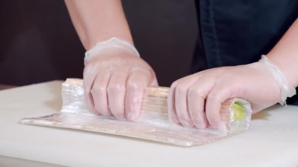 Primo piano di mani di chef maschile in guanti preparare deliziosi involtini di sushi con avocado in cucina. — Video Stock