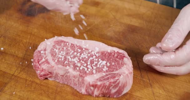 Крупным планом сочный мраморный бифштекс с грубой солью и специями на столе. Процесс приготовления мяса — стоковое видео