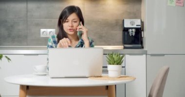 Cep telefonuyla konuşan genç bir kadın ev ofisinde dizüstü bilgisayarla çalışıyor. Kadın işçi, kablosuz cihazları kullanarak müşteriye profesyonel tavsiyeler veriyor. Uzak çalışma.