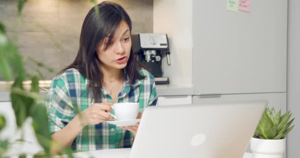 Mujer joven bebiendo café y hablando en video comunicación con colegas o amigos utilizando el ordenador portátil en casa. Trabajo remoto o concepto de educación en línea — Vídeos de Stock