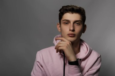 Genç ve çekici bir adamın portresi, güçlü bir şekilde belirgin elmacık kemikleri, model görünüşü, kahverengi saçlı, pembe kapüşonlu gri eğimli arka planda bir sandalyede oturuyor.
