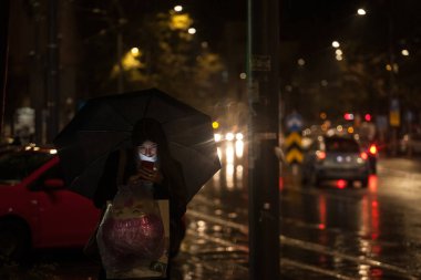 BELGRAD, SERBIA - 7 Ekim 2020: Coronavirus covid 19 sağlık krizi sırasında, yağmurun altında akıllı telefonundaki uygulamaları kontrol eden yüz maskesi takan genç kadın