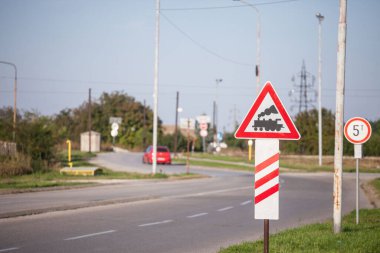Typical European railroad crossing sign on a rural road indicating an unguarded level crossing soon, with its iconic black steam locomotive on a red triangle. clipart
