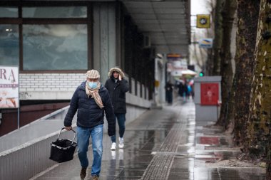 BELGRAD, SERBIA - 3 Nisan 2021: Coronavirus covid 19 sağlık krizi sırasında, Belgrad caddesinde yağmurun altında elinde alışveriş çantasıyla yürüyen solunum maskesi takan yaşlı adam
