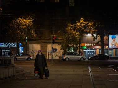 BELGRAD, SERBIA - NOVEMBER 15, 2020: Coronavirus covid 19 krizi sırasında gece elinde alışveriş çantası ve alışveriş arabasıyla solunum maskesi takan yaşlı bir kadının seçici bulanıklığı