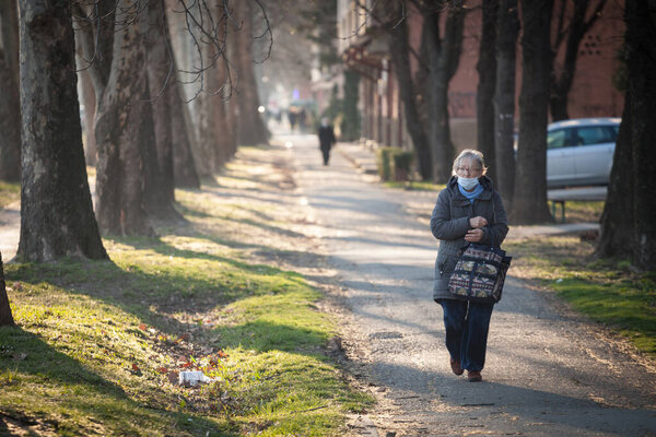 ПАНЦЕВО, СЕРБИЯ - 7 марта 2021 года: Старуха, идущая зимой по улицам Панчево в маске для лица, держащая сумку с продуктами во время кризиса Coronavirus Covid 19