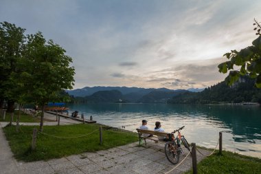BLED, SLOVENIA - 12 Haziran 2021: Blejsko Jezero şatosu Blejski Hrad ile birlikte Bled Gölü 'nün Panorama' sı, yaz günbatımında bisikletlerin yanında oturan gençlerle birlikte. Bled Castle büyük bir anıttır.