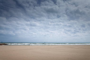 Valencia 'nın geniş bir sahili, kumlu bir plajı, dalgaları ve bulutlu bir gökyüzünün altında yelkenlileri var. La Malvarrosa 'daki Akdeniz kıyı manzarası sakin, sezon dışı havasında..