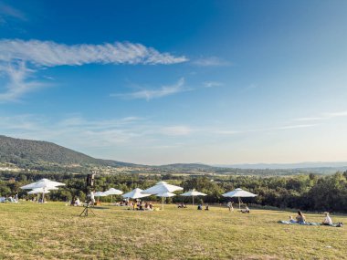 KOSMAJ, SERBIA - 13 Eylül 2025: Kosmaj tepelerine doğru Kabine Bira Fabrikasının çimlerinden geniş görüş. Ziyaretçiler, kırsal bölgeler uzaklığa doğru genişledikçe beyaz şemsiyelerin altında battaniyeler üzerinde dinleniyorlar..