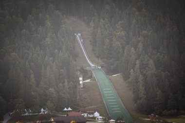 Slovenya 'nın Ljubno ob Savinji kentindeki kayakla atlama tepesinin seçici bulanık manzarası yazın yukarıdan görülüyor. Girişi, kalkışı ve yeşil iniş yamacı ormanı, bir yarışma ve eğitim yerini kesiyor..