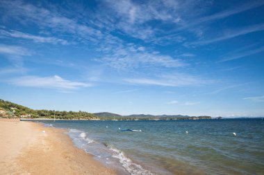 Akdeniz 'deki La Londe les Maures' daki Plage de Miramar plajı boyunca Panorama. Fransız Rivierası 'nda açık bir gökyüzünün altında Hyeres Körfezi boyunca kayıklar ve alçak tepeler ile geniş kumlu bir kıyı şeridi kıvrılır..