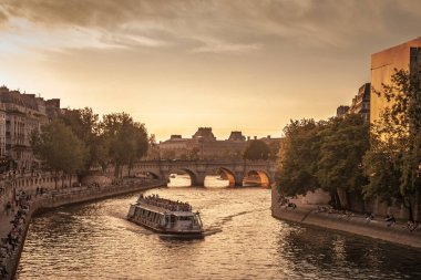 PARIS, FRANCE - 22 Haziran 2025: Paris 'teki Quai des Orfevres' in yanındaki Seine nehrinde Pont Neuf köprüsü ve Ile de la Cite 'de ılık günbatımı ışığında bir Bateau Mouche dolaşır. Gezginler nehir kıyısında sıralanır..