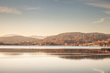 Klagenfurt, Avusturya 'daki Worthersee Gölü' nün sakin bir günbatımında panoramik manzarası. Görüntü parıldayan suyu, uzak tepeleri ve yumuşak ışıkla aydınlık gökyüzünü gösteriyor..