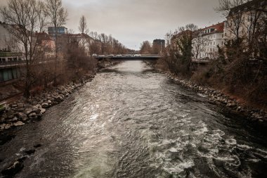 Avusturya 'nın Graz kentindeki Mur nehrinin panoramik manzarası, akan sular, kayalık bankalar ve çıplak kış ağaçları, sakin gri gökyüzü altında kentsel mahalleleri ve nehir kenarındaki binaları birbirine bağlayan bir köprü.