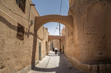 YAZD, İRAN - 10 AĞUSTOS 2016: İran 'ın Yazd kentindeki eski dar bir dar sokakta, çamur tuğlalı duvarlar ve güçlü güneş ışığı ve gölgelerle yürüyen yaşlı adam, İran' daki günlük yaşamında.