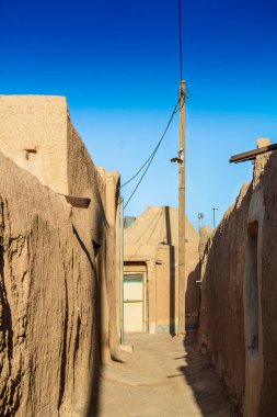 İran 'ın Yazd kenti yakınlarındaki bir çöl köyüne uzanan çamurdan duvarlarla kaplı dar bir ara sokak, açık mavi gökyüzünün altında geleneksel kerpiç evleri, elektrik direkleri ve telleri ve sıcak güneş ışığını gösteriyor..
