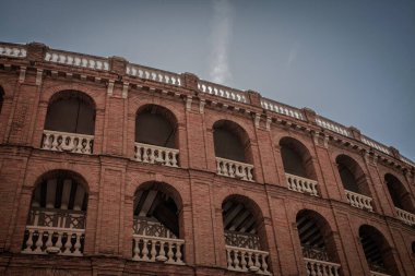 Valencia, Valencia 'da kırmızı tuğlalı bir arena olan Plaza de Toros de Valencia' nın alçak açılı görüntüsü bulutlu gökyüzünün altında kemerli galeriler ve beyaz balyalarla tarihi bir boğa güreşi simgesini vurguluyor..