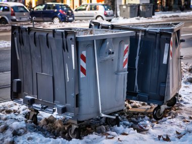 Büyük tekerlekli çöp konteynırları, alacakaranlıkta bir şehir caddesinde, buzlu kaldırım ve kış kalıntılarıyla sulu kar içinde, belediye atığı toplama, temizlik işleri ve soğuk hava koşullarını gösteren..