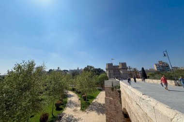 VALENCIA, İspanya - 6 Nisan 2025: Serranos Kuleleri (Torres de Serranos) Valensiya 'nın Pont dels Serrans kentinden yayalar ortaçağ şehir kapısına ve günışığı altındaki tarihi kalelere doğru ilerliyorlar.
