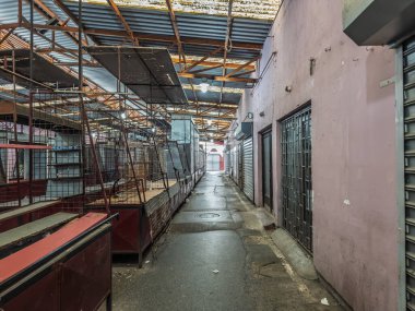 Sırbistan 'ın Subotica pazarında kapalı büfeler, kablo örgüsü tezgahları ve metal çatı yapısını gösteren sessiz bir kapalı sokak. Islak zemin ve floresan ışıkları mesai saatleri dışında atmosfer yaratır..