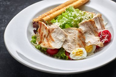 Tavuklu Sezar salatası, marul, domates, peynir ve yumurta, beyaz tabakta, siyah arka planda.
