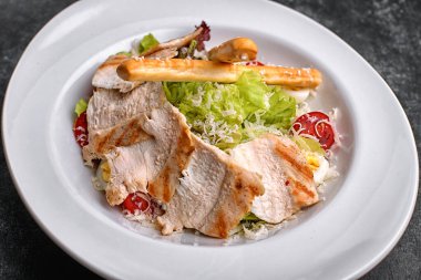 Tavuklu Sezar salatası, marul, domates, peynir ve yumurta, beyaz tabakta, siyah arka planda.