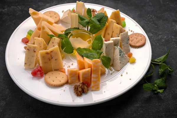 Cheese plate, assorted cheeses with mint, candied fruit, honey and cookies, on a white plate, on a dark background