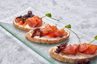 Üç bruschetta, alabalık, somon, krem peynir ve mikroyeşil beyaz arka planda cam bir tabakta.