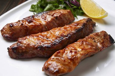 Üç parça ızgara somon fileto, alabalık, otlar ve limonla, beyaz bir tabakta, ahşap bir arka planda.