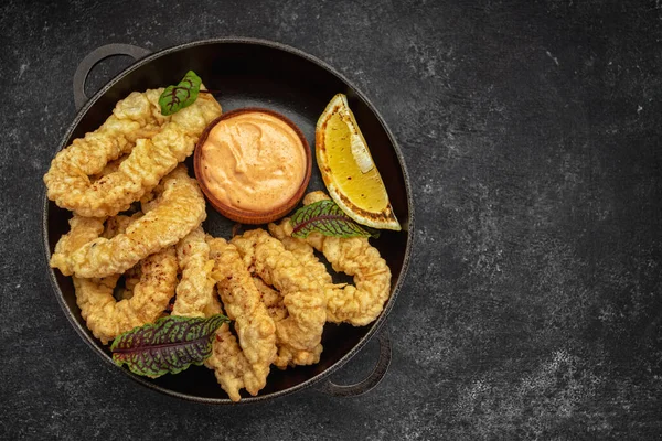 Anillos dorados de patatas fritas de calamar en una sartén redonda con salsa, hierbas y limón sobre un fondo oscuro sin decoraciones —  Fotos de Stock