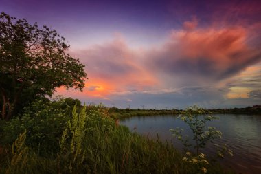 Gölün kenarında muhteşem bir gün doğumu. Önünde renkli bulutlar ve bitkiler var.