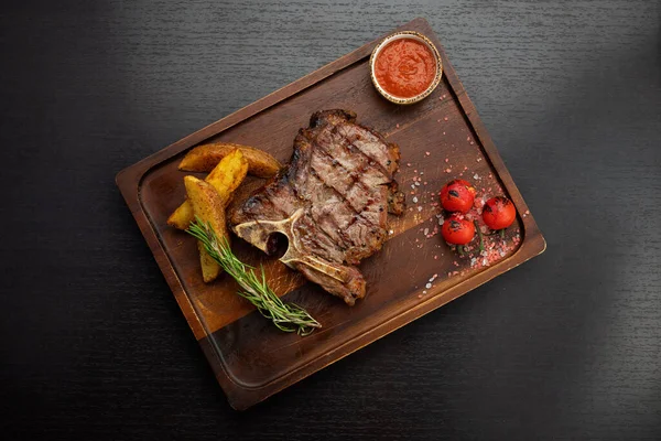 T-bone steak fából készült asztalon burgonyával, szósszal, cseresznyeválasztóval, sötét asztalon. — Stock Fotó