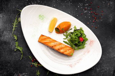 Kızarmış somon bifteği, şifalı otlar ve beyaz bir tabakta koyu bir arka planda limon.