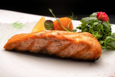 Kızarmış somon bifteği, şifalı otlar ve beyaz bir tabakta koyu bir arka planda limon.