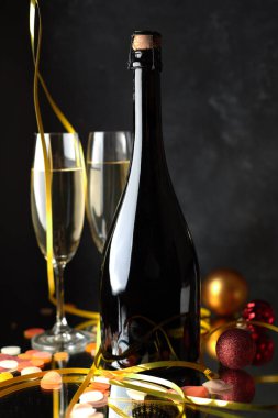 Şampanya şişesi, bardaklar ve koyu arkaplanda Noel süslemeleri olan zarif bir kompozisyon. Yeni yıl ve bayram teması için mükemmel..