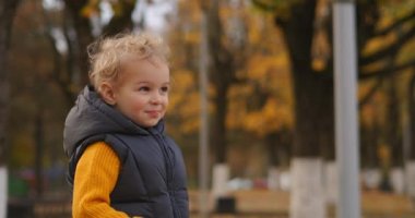 Sonbahar parkında sarı kıvırcık saçlı küçük çocuk, orta boy gülümseyen çocuk portresi.