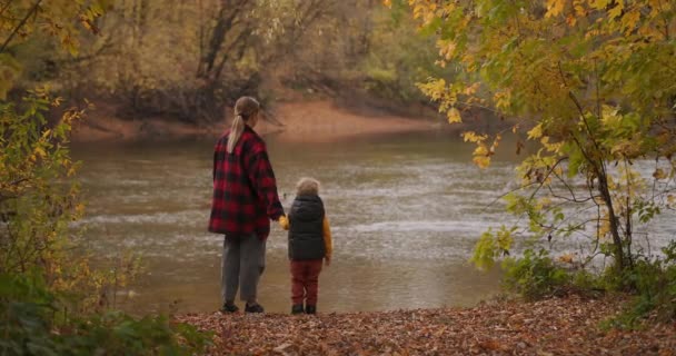 Vrouw met haar kleine kind in de natuur, mensen staan aan de kust van pittoreske bosvijver op herfstdag — Stockvideo