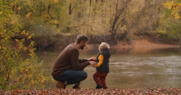 Szczęśliwy ojciec rozmawia z synem w lesie, stojąc na wybrzeżu jeziora lub rzeki w jesienny weekend, wycieczka rodzinna — Wideo stockowe